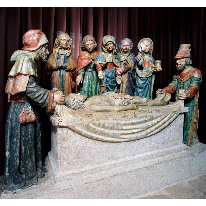 Source caption: "Bordant l’église, le cimetière possède une chapelle qui renferme une remarquable Mise au Tombeau groupant six personnages autour du Christ étendu sur le linceul dont Nicodème et Joseph d’Arimathie soulèvent les extrémités. Derrière le tombeau se tient la Vierge, douloureuse et recueillie, entourée de saint Jean l’Evangéliste et des « trois Marie », Cléophas, Salomé et Madeleine reconnaissable à son vase d’aromates."