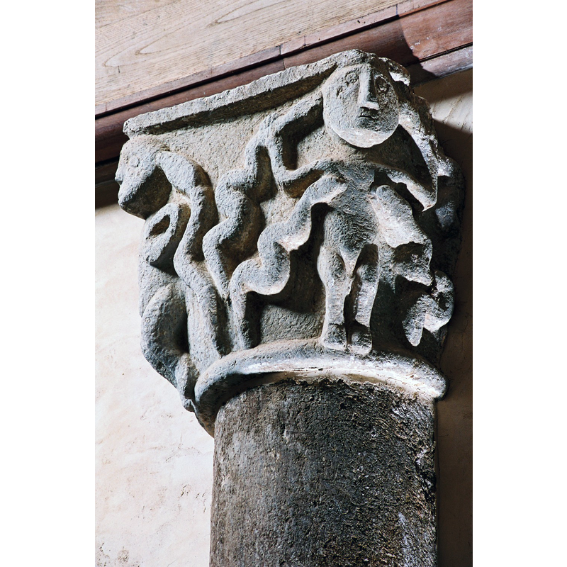 Source caption: "A l’intérieur, des demi-colonnes reposant sur des consoles portent des chapiteaux, injustement méconnus. L’un d’eux représente des atlantes, mains accoudés sur les hanches, traduisant bien l’effort nécessaire pour supporter de tailloir. Un autre représente la luxure aux mamelles pendantes, dévorée par des serpents."