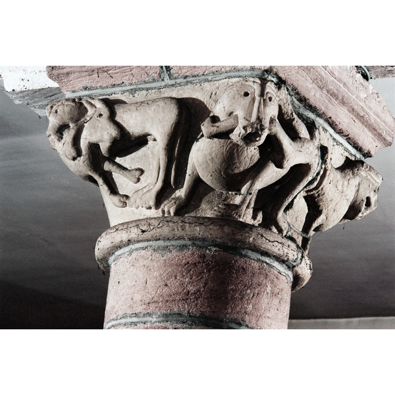 Source caption: "A l'intérieur, un chapiteau présente [...] une composition singulière : une tête de lion monumentale, à l’angle de la corbeille, domine un homme accroupi dans une position acrobatique."