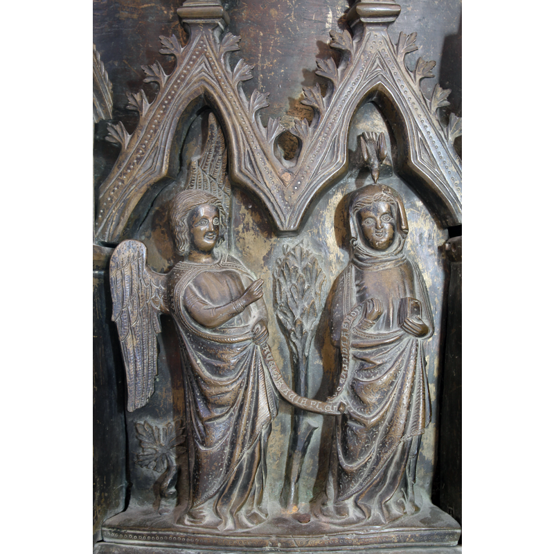 Gabriel has his right hand raised in benediction; his left hand holds one end of an inscribed scroll, the other end held in Mary's tight hand; a closed book in Mary's left hand; a bird [Holy Ghost?] in a trefoiled arch-head above Mary points its beak down at her head; a large bush or small tree between the two figures, in the background; a plant at Gabriel's left side, by his feet