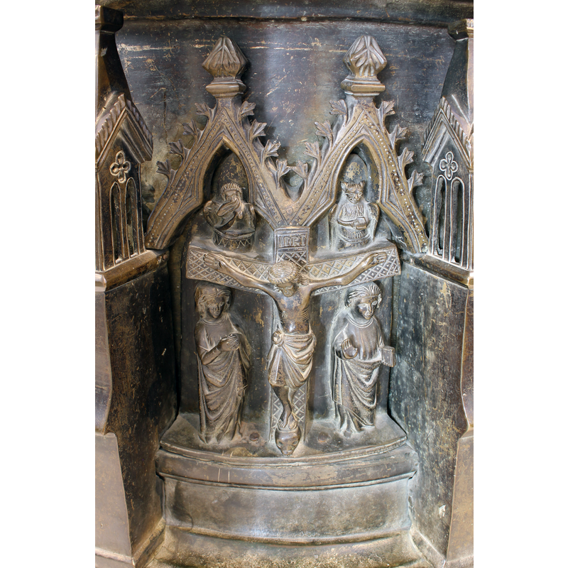 Mary, on the left, hands together in prayer; John, on the right, right hand with palm out, left hand holding a closed book; both of them face the viewer; the surface of the cross is patterned; the upper end bears the "INRI" sign; s human skull beneath Christ's feet; the trefoiled arch-heads atop house two winged figures; the one on the left appears human; the one on the right appears feline