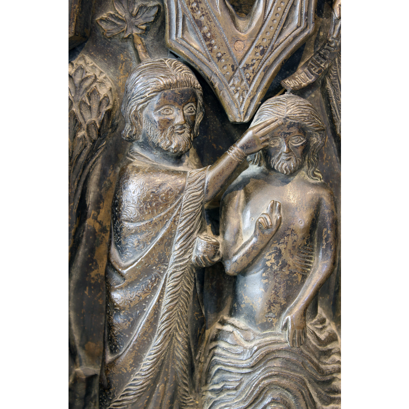 John carries a small container in his left hand, his right hand in the common benediction manner touches Chirst's forehead; the Manus Dei up in the right side arch-head is accompanied by an inscribed scroll