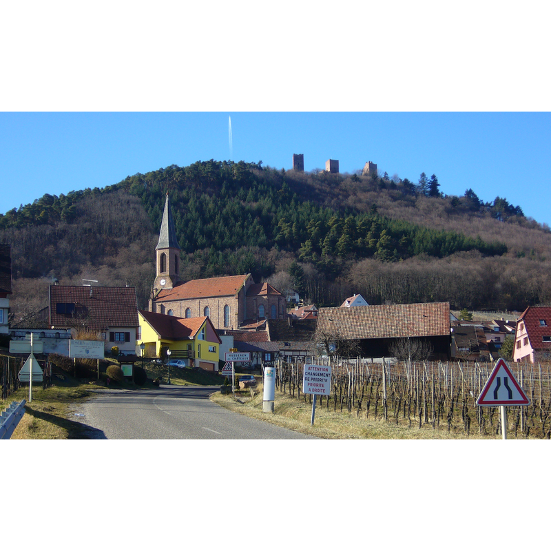 Source caption: "L'entrée du village de Husseren-Les-Châteaux (Haut-Rhin)" -- showing a view in the background, on the Schlossberg hill, of 'Les Trois Châteaux d'Eguisheim' [i.e., Wahlenbourg, Weckmund and Dagsbourg]