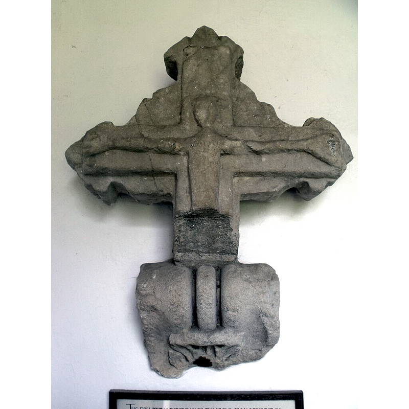 Source caption: "Stone crucifix, St Peter & Paul's church, Langton by Spilsby. The following reference to the cross appeared in “Lincolnshire Notes and Queries” Vol VII Page 30 (1902): “An interesting discovery has recently been made by Dr Cochrane, the Rector of the Parish: In a plantation adjoining the Rectory grounds has been found a large portion of an ancient stone crucifix, with the Saviour outstretched on the Cross, perfect down to the waist: the lower part having not yet been found, as it is to be hoped it may be. The style of the Cross, with rude trefoil finials to the top and two arms, and the carving of the figure, are so primitive, this it is probably a Saxon relic. The relic bears some resemblance to a very old carving of the Crucified Saviour, still to be seen, cut in the natural rock of a cave in the side of a hill named Carcliff Tor, near Rowsley in Derbyshire, a retreat where secret worship was probably conducted by early Christian converts.”"