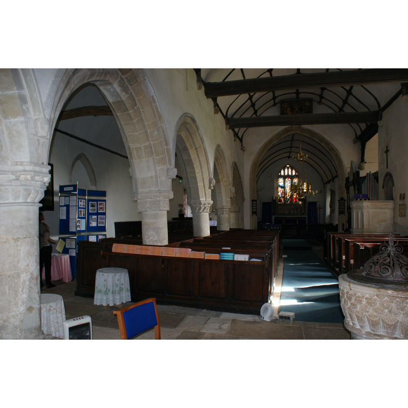 Source caption: "The 14th Century (Decorated) nave and north aisle (remodelled in 1460) of the Century Church of St. Mary's Burton, Nettleton, 03/14. Some sources say the arches date from 1460 and placed on restored 12th Century Norman columns, but other sources state the columns also date from 1460 and are merely copies. Note the arches and columns lean due to subsidence at some distant time in the past. View is towards the chancel." -- the old font is partially visible in the bottom-right corner of the image