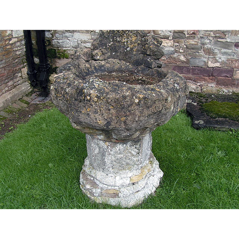 this object is a composite font; the basin is indeed what remained when the 15thC font was damaged, but the pedestal  is not the original; the remains of the old base are kept inside the church, by the side of the 19thC font in use