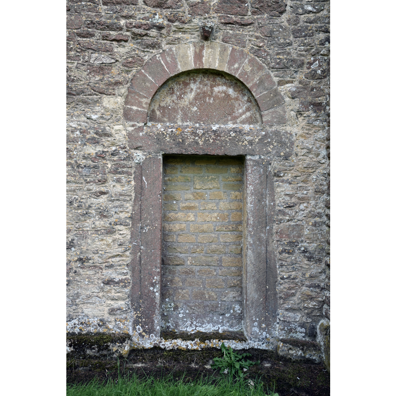 Source caption: "Winstone Church (St. Bartholomew), Gloucestershire, 6 September 2016. Mid 11th Century Saxon/Norman 'overlap' church with 15th Century saddleback tower in similer [similar?] style, restored in 1876 by Waller & Son. Pictured is the blocked north doorway with Saxon style inclined jambs and Norman style round arch with plain tympanum."