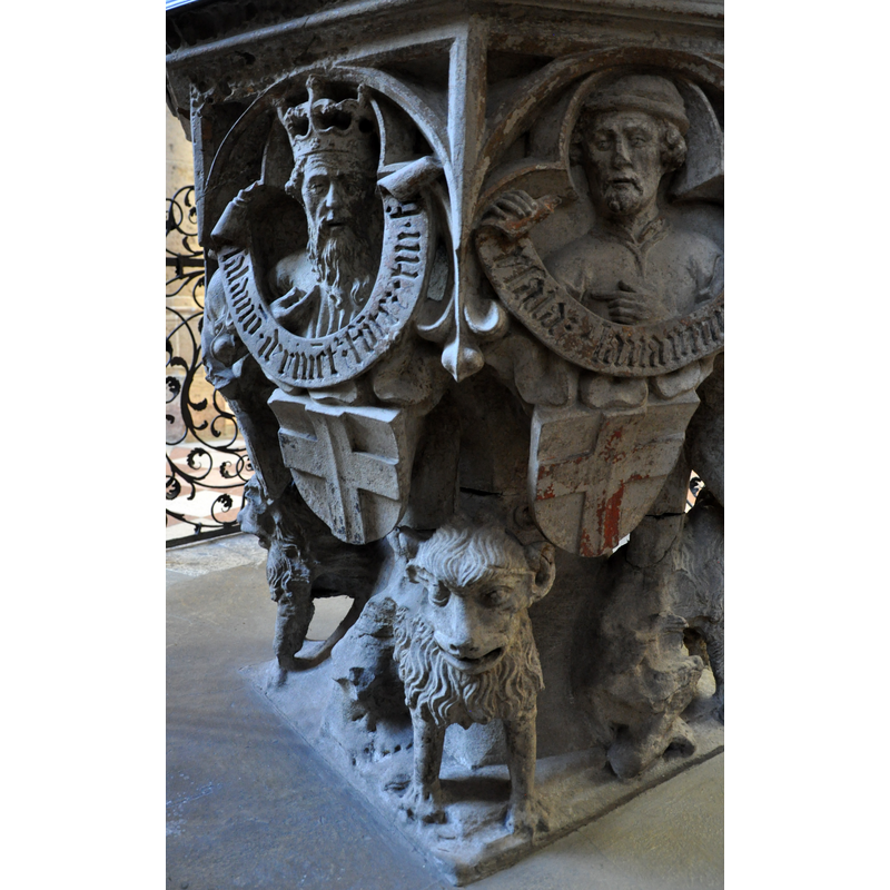 on the left side of the image -- Source caption: "Ulm, Ulmer Münster, Taufstein (1474), Detail: Figur mit dem Wappen des Kurfürsten von Köln (links), rechts mit dem Wappen des Kurfürsten von Trier".