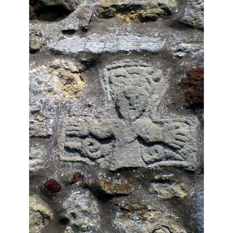 Source caption: "Ancient stone in south wall of Sinnington church. A tour around the outside and inside of All Saints church reveals several ancient stones dating back to the old Norman church which stood here previously. This appears to be the remains of a cross head showing Christ on the cross and may well be of Saxon origin."