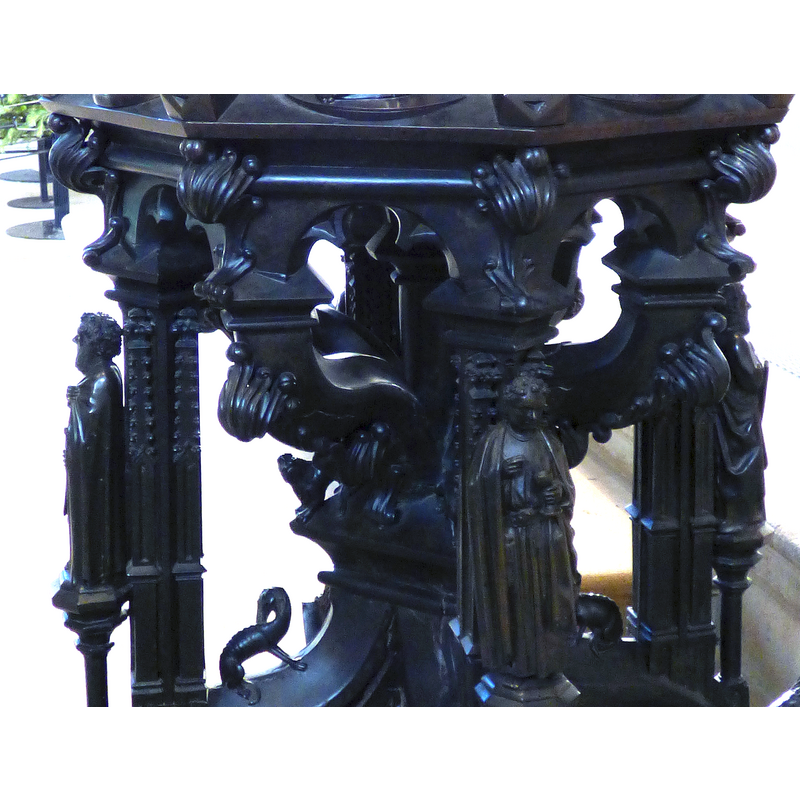 raised on tall platforms on the outer side of the four outer colonnettes of the base; like to figures on the basin sides, these appear to hold objects in their hands -- the figure in the centre of the image appears to be a cleric holding a chalice in his left hand and blessing it with his right hand