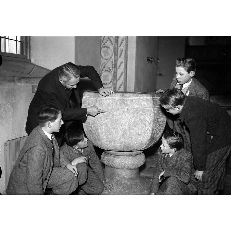 Source caption: "Folkskollärare Hugo Fredriksson visar dopfunten i Alunda kyrka för elever. Eleven längst till vänster troligen Malte Mårtensson. Bilden ingår i "Olands kommuns bildarkiv" inköpt 1999 från fotograf Bo Larsson, Alunda."