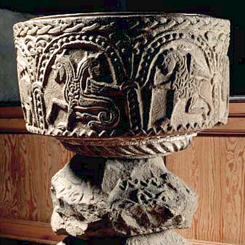only a small section has survived, seen here on the right, below the zig-zag fragment -- the original base was probably of the two-tiered type, with broad rope-patterned mouldings and heads at 90-degree angles on both levels as well; it is now little more than a stump, to which was added a later lower base to raise the height of the font to a functional level