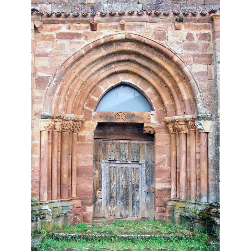 Described in Arteguias [www.arteguias.com/monasterio/sanmillanlara.htm] [accessed 13 June 2020]: "La portada occidental pertenece a otro momento distinto y posterior ya que, tanto su estructura como escultura denotan gran diferencia con la anterior. Tiene cuatro arquivoltas apuntadas y capiteles con bestias silenses como arpías, varios temas figurados muy deteriorados (quizás uno de ellos trate de la degollación de San Juan Bautista), dos grandes rostros humanos y una representación cinegética de montería del jabalí. Queda el dintel -con cruz patada- de un tímpano desaparecido y una de las mochetas con cabeza leonina."