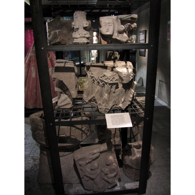 Source caption: "Olika kapitäl och valvkonsoler från Skara domkyrka på Västergötlands museum. De återanvändes inte vid Helgo Zettervalls restaurering 1886-1894 på grund av skador på dem. Därför deponerades de istället i museet. För att ersätta dem höggs nya kapitäl under restaureringen." [NB: the caption does not mention the font seen here in the bottom left corner, but refers to the other stones as a mixed variety from the earlier church not re-used in the 19thC restoration by Helgo Zettervall, all of them shown in the storage fecility at the Västergötlands museum -- the relief partially visible in the bottom-right corner appears to be the offerings by Cain and Abel]