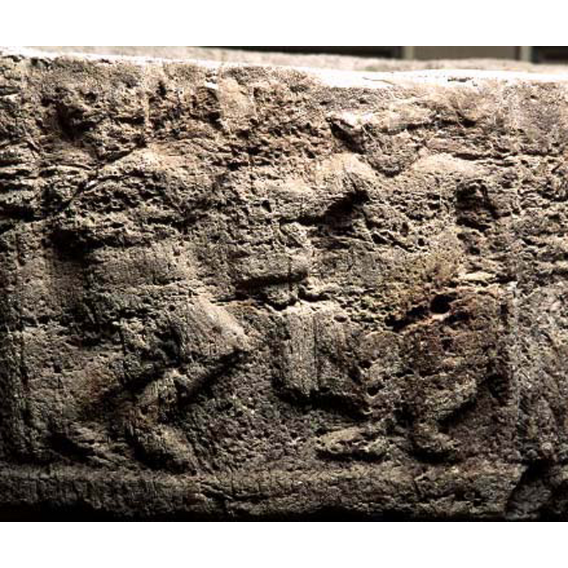 three human figures are still discernible in the badly-eroded remains of the carving: on the right is a seated figure facing the viewer; on the left is a standing figure facing the seated figure on the right; a much smaller figure, of whom only part of the body and two tiny feet stand on the knee of the seated figure facing it; the arms of the left fugure and one arm of the right figure appear to be holding the 'child'