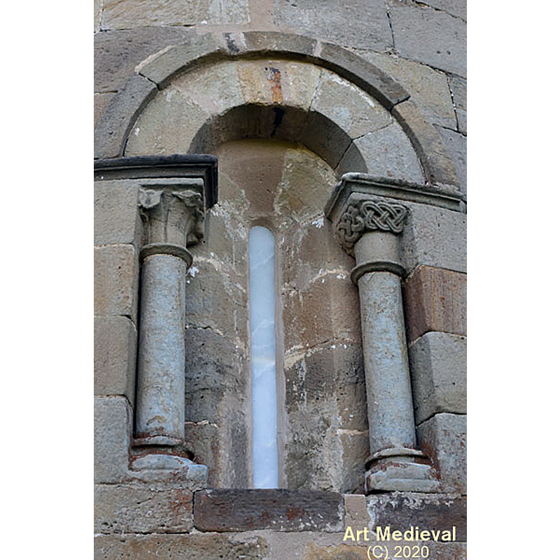 Source caption: " En el tramo central del ábside se abre una ventana de medio punto y doble derrame. El arco está rodeado por un sencillo guardapolvo liso y se apoya en dos columnas con los capiteles esculpidos. El del lado izquierdo es de tipo vegetal, mientras que el del lado derecho tiene un entrelazado en la parte superior."