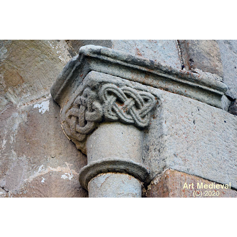 Source caption: " En el tramo central del ábside se abre una ventana de medio punto y doble derrame. El arco está rodeado por un sencillo guardapolvo liso y se apoya en dos columnas con los capiteles esculpidos. El del lado izquierdo es de tipo vegetal, mientras que el del lado derecho tiene un entrelazado en la parte superior."