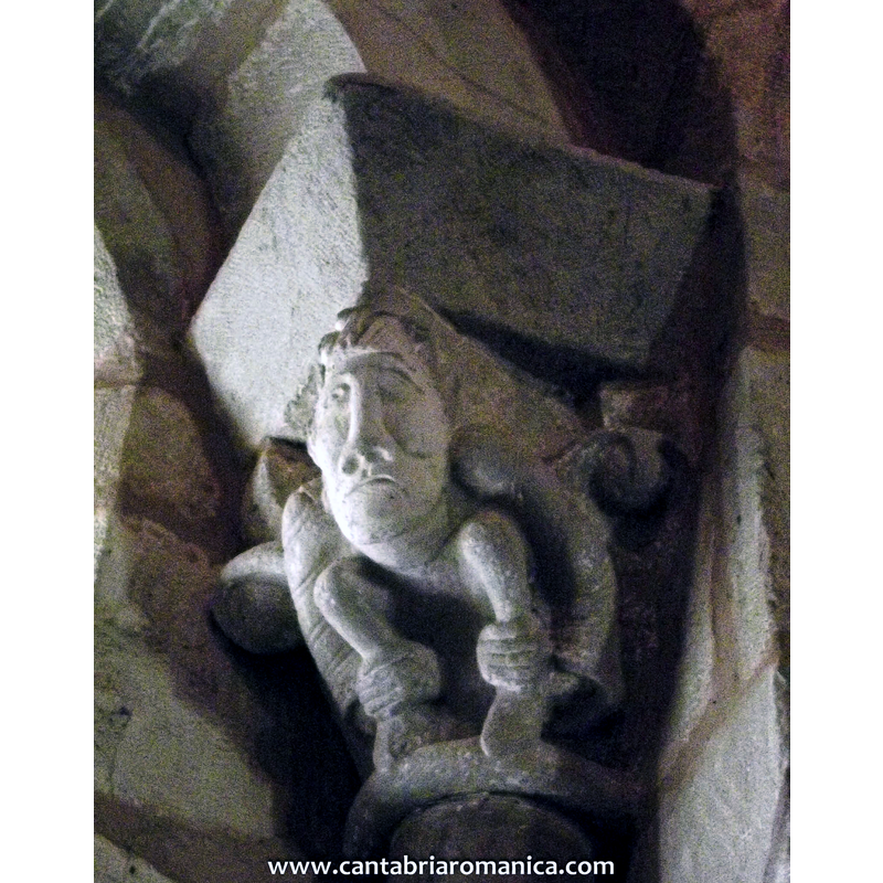 Source caption: "En el centro una ventana simple de arco doblado sin arquivoltas. Los capiteles de esta ventana son una de las cosas más interesantes de la iglesia de San Juan Bautista. El izquierdo se decora con un cimacio angular muy parecido a los que podemos encontrar en Cervatos, y decoración  vegetal bastante deteriorada. Al respecto del capitel derecho, con cimacio biselado y liso, tiene un alto interés en lo referente a su iconografía ya que representa la figura de un hombre vestido con una túnica  y en cuclillas lo que muchos expertos han determinado como la actitud de un hombre que está defecando."