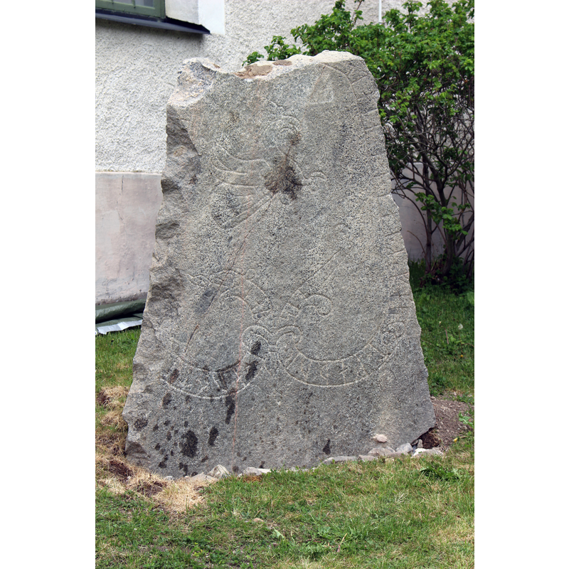 Source caption: "Runstenen Upplands runinskrifter 874" -- Descriprion in Fornsök Riksantikvarieämbetet: "Runsten, U 874, ca 1,5 m h, 1,32 m br och 0,5 m tj vid roten, avsmalnar uppåt. Stenens fulla längd är 1,92 m . Tolkning: Jarl och ... stenen efter Gerfast, sin fader. Låg tidigare som tröskelsten i vapenhuset till den medeltida kyrkan. Runstenen återfanns 2016-10-03 av Emelie Sunding, Upplandsmuseet i samband med en schaktning för ny ringledare runt kyrkan. Den restes på nuvarande plats 24 maj 2017, ca 9 m NV om fyndplatsen.(Besiktning 2017, lst dnr 435-2588-17)" [NB: the Runic stone had been used earlier as a threshold stone in the porch [vapenhus] of the church]
