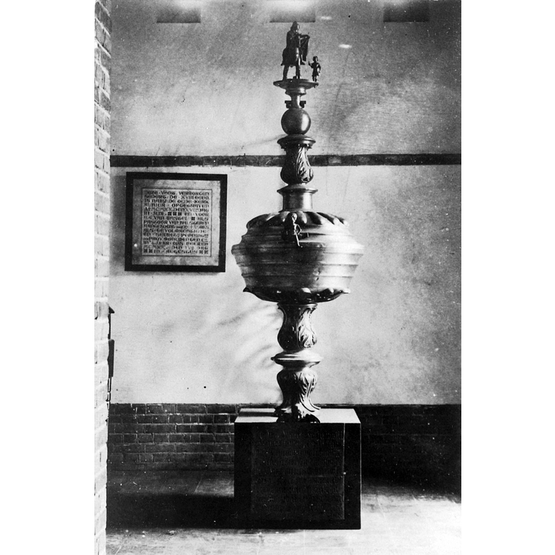 Source caption: "De doopvont stond oorspronkelijk in de ruimte onder de noordelijke toren, waar thans de Mariakapel is ondergebracht. Het voetstuk herbergt de koperen gedenkplaat waarop de reden van de schenking is vermeld." [NB: the box-shaped platform on which the font stands would later be replaced by a modern round stone plinth decorated with a gadrooned pattern after the one on the metal font cover]