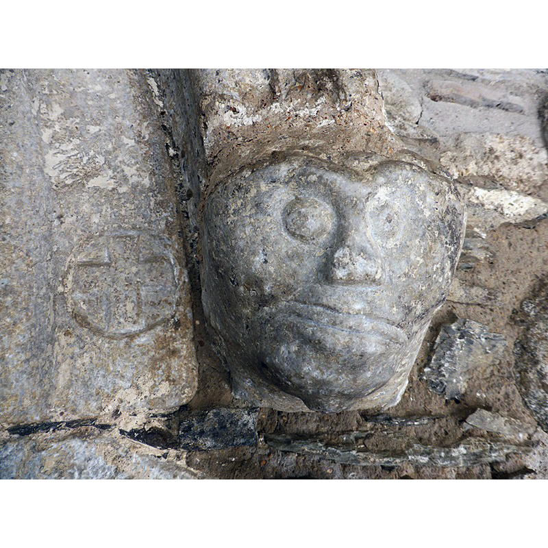 Source caption: "Portada a poniente, bajo un pórtico de época moderna, formada por tres arquivoltas en bocel, que llegan al suelo, enmarcadas por un guardapolvo adornado con cinco cabezas, las dos de su base a modo de ménsulas. Las arquivoltas muestran cruces, estrellas y aspas grabadas."