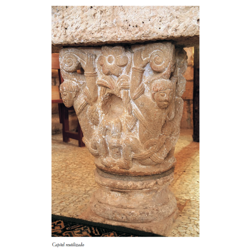 Source caption: "Reutilizado como soporte de la mesa de altar encontramos un capitel románico de grandes dimensiones, buena calidad y afortunadamente bastante bien conservado, alzado sobre una basa de perfil ático y amplia escocia, ambas piezas procedentes de una columna entrega, quizá de la nave primitiva. La cesta se decora con dos personajes acuclillados que con sus brazos alzados se agarran a las grandes volutas que, entre florones, coronan el capitel. Estas figuras aparecen enredadas por tallos que son sujetados por otros dos personajes en los laterales de la pieza, mientras que en el frente estos tallos se anudan y dan lugar a piñas. Su buena factura recuerda modelos onienses y los rostros los de algunos canecillos de San Pedro de Tejada."