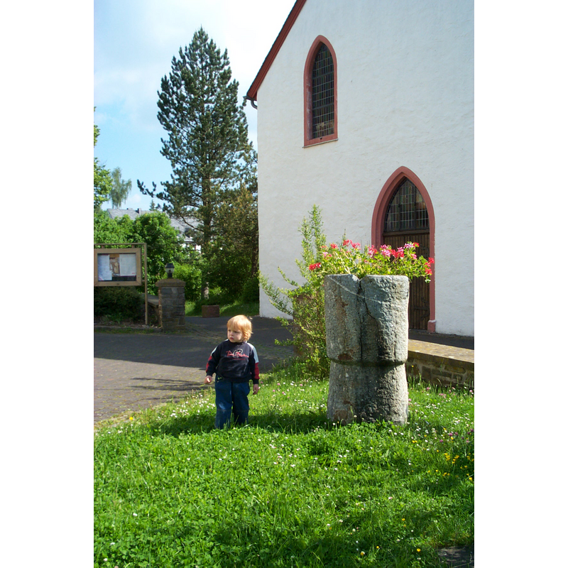 Source caption: "Romanischer Taufstein neben der Kirche in Binscheid, Gemeinde Üttfeld (Eifel, Deutschland). Der Sage nach hat der heilige Willibrord einen Taufstein aus einem heidnischen Menhir meißeln lassen und darin die ersten Christen getauft."