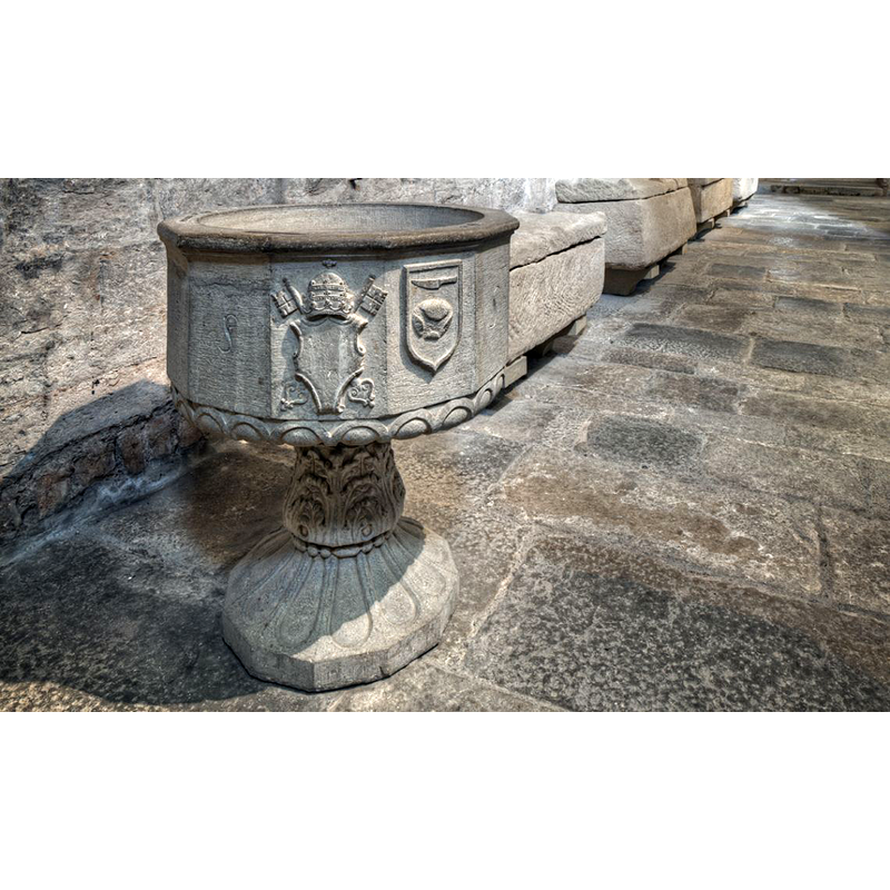 Source caption: "La iglesia fue parroquia del burgo de Sant Pere y, por tanto, se administraba la mayor parte de sacramentos. De aquí la existencia de esta pila bautismal, datada en 1550. La pila tiene doce caras, entre las que se encuentran dos escudos. Uno con el campo liso, ostenta la tiara y las llaves de san Pedro, por la advocación del monasterio, y el otro tiene en el campo un cuchillo y una piel, la herramienta y el producto del trabajo de los curtidores. El gremio de los curtidores, que tenían los talleres en el burgo, fueron los que financiaron la pila."