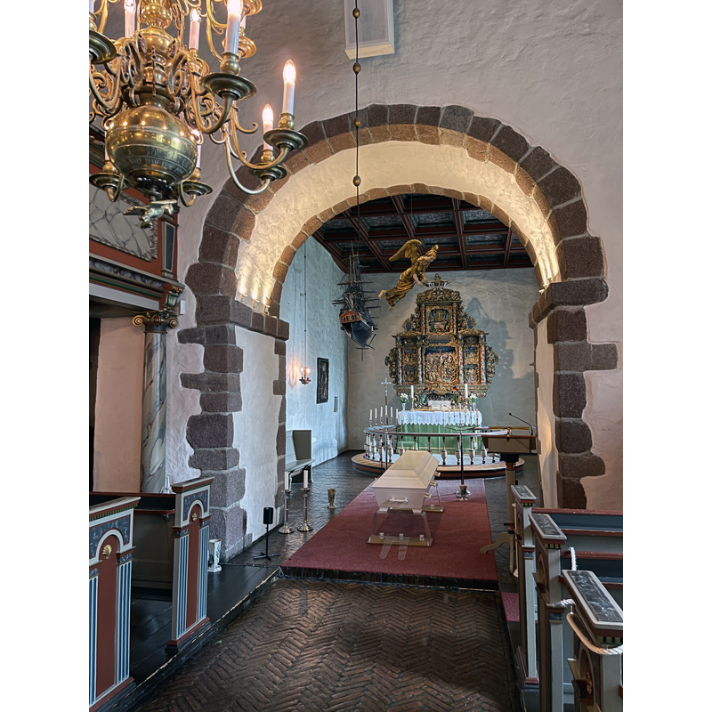Source caption: " Interiør i Borre kirke, en romansk middelalderkirke i Horten kommune i Vestfold og Telemark fylke:
    Alter med alterring og barokk altertavle (engelsk: altar piece) skåret av treskjærer Abel Schrøder d.y. 1665 og malt og forgylt i 1741
    Tak i koret dekorert med tekster, motiver og rosemaling-lignende ornamenter
    «Dåpsengel» (baptismal angel); dåpsfatet holdes av en svevende engel som fires ned fra taket
    Votivskip, skipsmodell, «kirkeskip»
    Kiste (coffin) før en gravferdsseremoni
    Lysekroner (chandeliers)
    Søyler, paneler og annet med malt marmorering, etc."