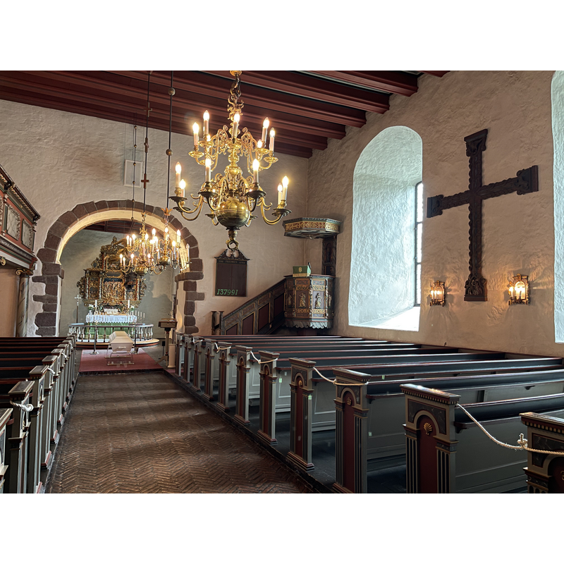 Source caption: " Interiør i Borre kirke, en romansk middelalderkirke i Horten kommune i Vestfold og Telemark fylke:
    Alter med alterring og barokk altertavle (engelsk: altar piece) skåret av treskjærer Abel Schrøder d.y. 1665 og malt og forgylt i 1741
    Tak i koret dekorert med tekster, motiver og rosemaling-lignende ornamenter
    «Dåpsengel» (baptismal angel); dåpsfatet holdes av en svevende engel som fires ned fra taket
    Votivskip, skipsmodell, «kirkeskip»
    Kiste (coffin) før en gravferdsseremoni
    Prekestol (pulpit) i renessansestil fra circa 1600, med billedframstillinger av dydene
    Salmetavle (hymn board)
    Lysekroner (chandeliers)
    Kirkebenker, benekerader (wooden pews, church benches)
    Søyler, paneler og annet med malt marmorering
    Kopi av Borrekorset (The Borre Cross) fra slutten av 1200-tallet, etc." -- the cross on the south wall is a replica copy; the original is in the KHM, Oslo