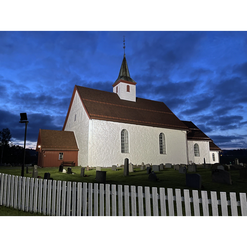 Source caption: " Ramnes kirke, en hvitkalket steinkirke fra middelalderen i Tønsberg kommune i Vestfold og Telemark fylke. Kirkegård/gravlund innrammet av hvitt stakittgjerde og med gravsteiner/gravstøtter/gravminner utenfor kirkebygningen. Ramnesveien passerer forbi rett utenfor kirkegårdsporten."