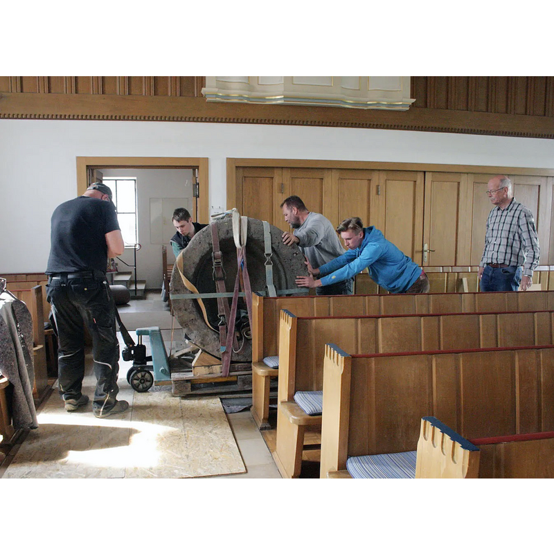 Source caption: "Steinmetzmeister Jörg Fichtner (grauer Pullover) und sein Team hatten schwer zu schleppen, als sie in dieser Woche den mittelalterlichen Taufstein in der Kirche im Hüttenberger Ortsteil Vollnkirchen aufstellten. Mit Hubwagen, Gerüst und Flaschenzug wurde der Taufstein an seinen Platz neben dem Altar gehievt. Im karierten Hemd: Frieder Heinze, Baubeauftragter der Kirchengemeinde für den Taufstein."