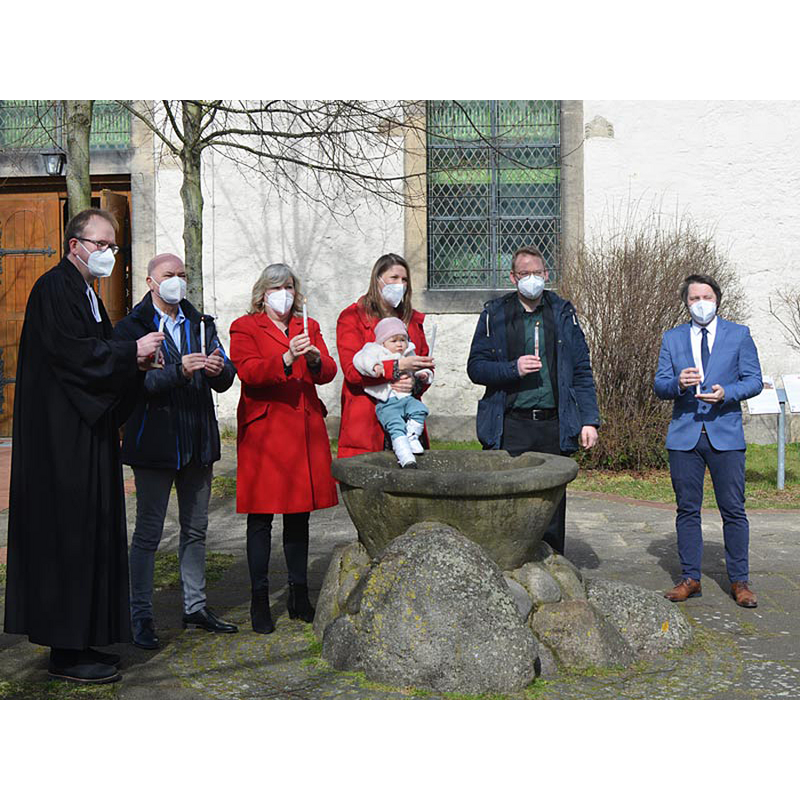 Source caption: "Pastor Rainer Müller-Jödicke (von links), Wolfgang und Gabriela Kasten, Jana, Mila Marie und Benjamin Buske und Diakon Merlin Langrehr geben das Osterlicht am Taufstein vor der Engelbosteler Martinskirche weiter . Foto: Andrea Hesse"