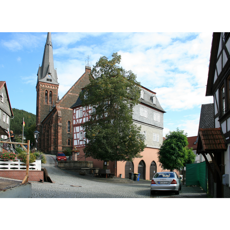 Source caption: "Der alte Marktplatz von Biedenkopf in der Altstadt (heute "Oberstädter Marktplatz", "Oberer Markt" oder "Oberer Marktplatz" genannt). Blick (v.l.n.r.) auf die evangelische Stadtkirche, den Oberstädter Marktbrunnen, die Oberstadt-Linde und das erste Rathaus der Stadt; zwei weitere existieren noch. Fachwerkbau mit Mansarddach; 1717 abgebrannt, von 1732 bis 1935 Sitz der Stadtverwaltung. Zeitweilig Jugendherberge, nun Wohngebäude. Kellergeschoss mit gotischen Spitzbögen; inklusive Anbau (bis ca. Mitte 19. Jhdt. städtisches Brauhaus); Blick von der Hintergasse."