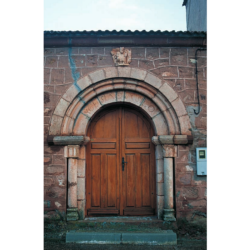 Source caption: "En el muro norte de la nave se encajó la primitiva portada románica, formada por un arco de medio punto y una arquivolta decorada con dos boceles que apoya sobre un par de columnillas con capiteles de hojas esquemáticas, como en Valvenedizo. Sobre ella se empotró un capitel en el que se representa a un obispo bendiciendo flanqueado por un acólito turiferario y otra figura mutilada."