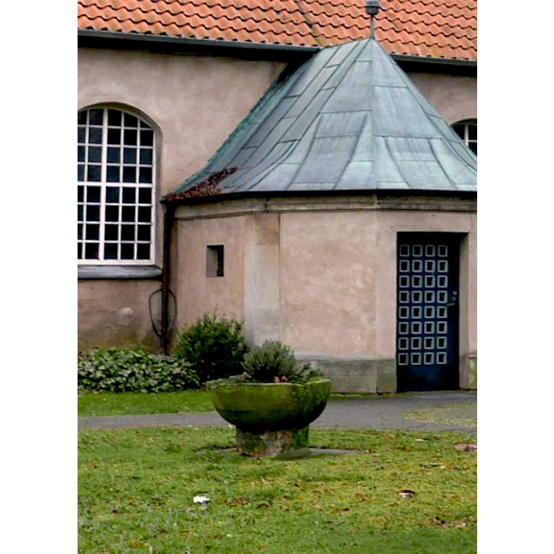 Source caption: "Auf eine mittelalterliche Steinplatte aufgesetztes rundes Sandsteinbecken, am oberen Rand Teile eines Bogenfrieses erhalten. Der romanische Taufstein der Basser Kirche wurde um 1100 gefertigt und ist heute als Blumenschale vor der nördlichen Vorhalle aufgestellt. Aufgrund seiner Bedeutung für die Orts-, Bau- und Kunstgeschichte sowie wegen der städtebaulichen Bedeutung mit prägendem Einfluss auf das räumliche Gefüge der Kirchenanlage von Basse besteht an seiner Erhaltung ein öffentliches Interesse."