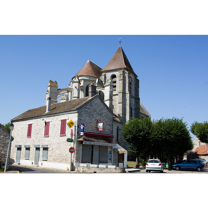 Source caption: "Eglise de Champcueil, Champcueil, Essonne, France" -- the apse and the northeast tower are partially visible here; the view is obstructed by the bar-tabac-brasserie 'Le P'tit Chancolia' in the foreground