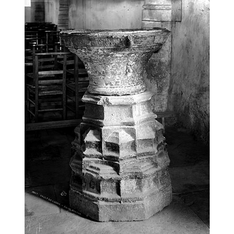 Source caption: "Bénitier en fonte de fer sur un socle en pierre. Sur une base à six pans ornée de moulures et de saillies dans le goût du 15e siècle, le bénitier en forme de cloche renversée décoré de filets en relief. A mi-hauteur, quatre têtes en saillie ; sur la plate bande supérieure d'où émergent deux anneaux, lettres gothiques IHS."