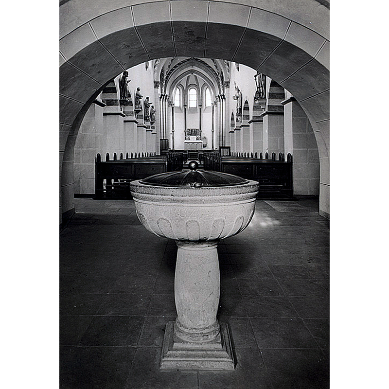 Source caption: "Taufbecken in der Pfarrkirche St. Pankratius Oberpleis. Dieses Bild zeigt den Taufstein, der bis 1974 unter dem Turm der Pfarrkirche St. Pankratius stand. Er wurde nach der Renovierung 1978 in der Krypta aufgestellt. An seine Stelle wurde der alte Taufstein aufgestellt, der bis dahin im Quadrum gestanden hatte. Dieser stammt ursprünglich aus Friesdorf und kam in den 1950er Jahren nach Oberpleis. Bevor er im Turm aufgestellt wurde, wurden die fehlenden Säulen, deren Ansätze man auf dem Bild im Quadrum gut erkennen kann, ersetzt (s. Link unten)."