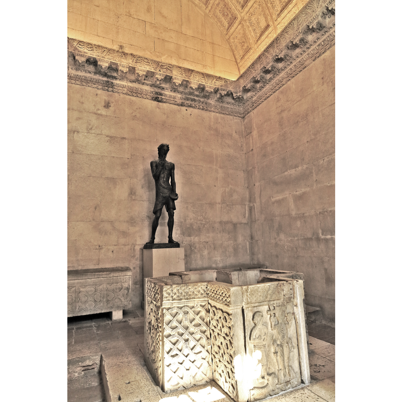Source caption: "Der Jupitertempel wurde nach der Christianisierung zur Taufkapelle und Johannes dem Täufer geweiht. Die Statue ist neuzeitlich, das Taufbecken stammt aus dem nachrömischen Mittelalter, gut zu sehen an den grob gehauenen Gesichtern."