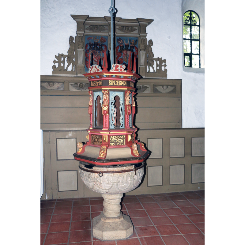 Source caption: "Romanischer Taufstein der St.-Marien-Magdalenen-Kirche (Erfde) mit Taufdeckel von 1635 aus der Werkstatt von Jürgen Heitmann dem Älteren, dahinter Gestühl mit dem Wappen von Herzog Adolf von Schleswig-Holstein-Gottorf und seiner Gemahlin (um 1570)."