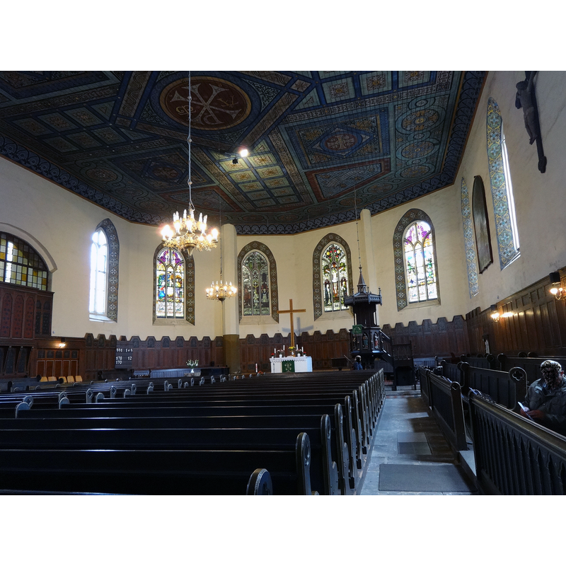 Source caption: "Die Stadtpfarrkirche St. Nikolai in Bad Liebenwerda ist ein spätgotisches Bauwerk, dessen genaues Baudatum unbekannt ist. Nach einem Brand setzte sich Friedrich III. (Sachsen) dafür ein, sie im Jahr 1515 wieder aufzubauen. Aus den Jahren 1519 und 1544 sind Besuche Martin Luthers überliefert. Im Innern befinden sich eine neugotische Kanzel sowie eine prachtvolle Flachdecke, die August Oetken im Jahr 1911 gestaltete. Die Glasmalereien in den vier Fenstern des Chors zeigen die Taufe Jesu, Geburt, Kreuzung und Auferstehung Jesu Christi. Sie stammen aus dem Jahr 1908 und entstanden in der Werkstatt von Gerstner und Werner in Görlitz und gehen auf Stiftungen des Apothekers Liebe sowie dem Büromaschinenhersteller Reiss zurück. Sehenswert ist weiterhin eine Fünte aus Sandstein aus dem Jahr 1671. sowie ein überlebensgroßes Kruzifix vor einem Triumphkreuz an der Südwand, das vermutlich aus dem 16. oder 17. Jahrhundert stammt." -- showing the top of the baptismal font behind the benches on the left, left of the altar, right of the psalms board, with a vase of flowers on top