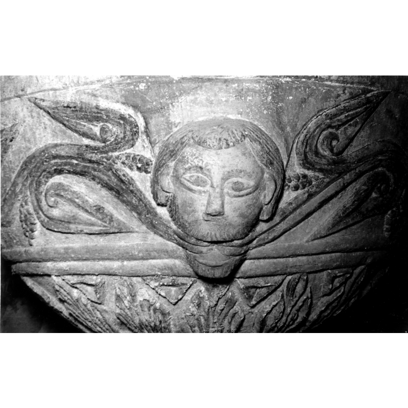 Source caption: "Detalle de la decoración de la taza de la pila bautismal; es una cabeza hum ana de ojos almendrados de cuya boca sale una guirnalda de hojas lanceoladas y r acimos que rodea la pila"; Fragmento de pila bautismal de la iglesia de Nuestra Señora de la Asunción"