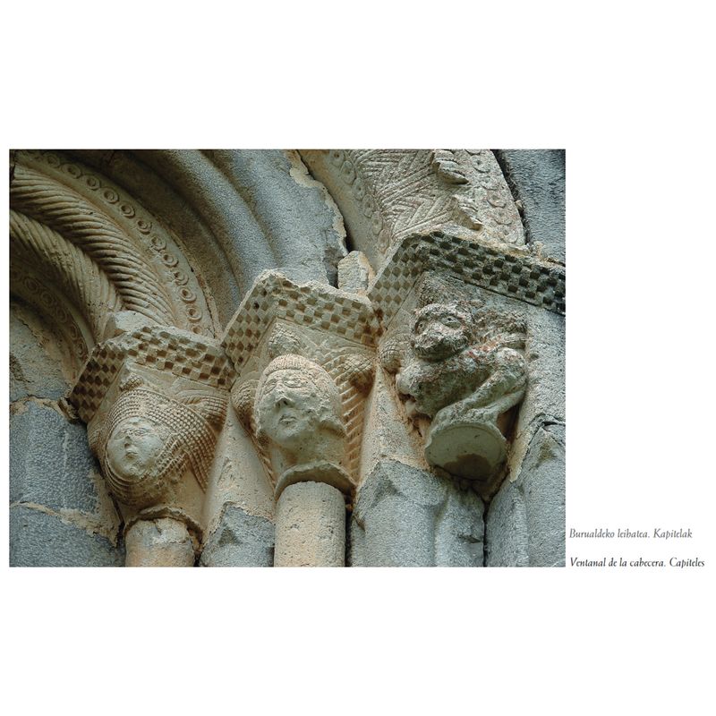 Source caption: "Los capiteles interiores muestran damas de estilizados rasgos y cubiertas con tocados de barbuquejo, decorados con incisiones que
forman siete bandas paralelas. En los capiteles centrales se representan rostros varoniles, enmarcados por cortas melenas, de ojos almendrados y facciones muy pronunciadas. En los dos exteriores se labra un tema animalista, muy repetido en el románico de Zigoitia y que transciende a otras zonas: el del carnicero con acusados rasgos de fiereza, mirando al frente con el cuerpo retorcido, y la cola elevada saliendo entre sus patas traseras".