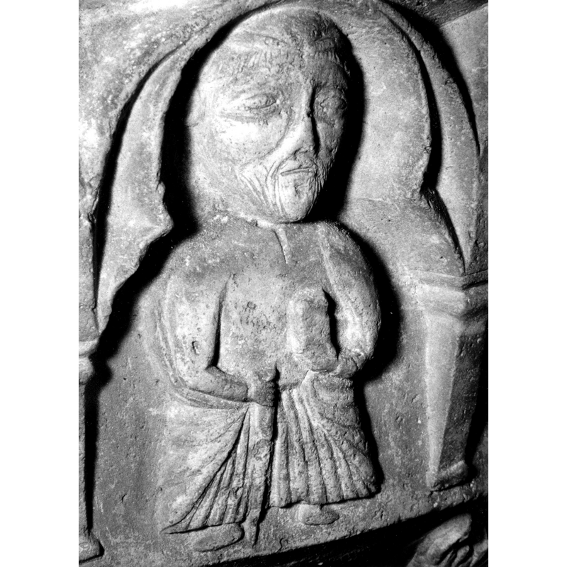 Source caption: "Detalle de una de las figuras de la parte superior de la pila bautismal gótica; se representa una figura de hombre, un apóstol, profeta o fraile franciscano con un libro en las manos, entre arquerías polilobuladas [...] pila bautismal de la iglesia de la Natividad de Hueto Arriba"
