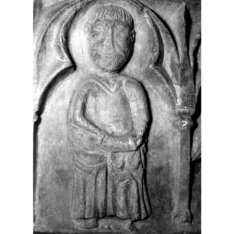 Source caption: "Detalle de una de las figuras de la parte superior de la pila bautismal gótica; se representa una figura de hombre, un apóstol, profeta o fraile franciscano con una cartela en las manos, entre arquerías polilobuladas [...] pila bautismal de la iglesia de la Natividad de Hueto Arriba"