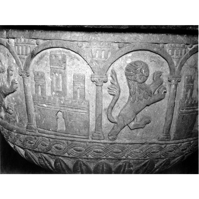 Aource caption: "Detalle de la copa de la pila bautismal; presenta una decoración de leones rampantes y castillos alternando cobijados por arquerías de medio punto sobre es beltas columnas. En las enjutas, construcciones almenadas [...] pila bautismal de la iglesia de la Asunción de Nuestra Señora"