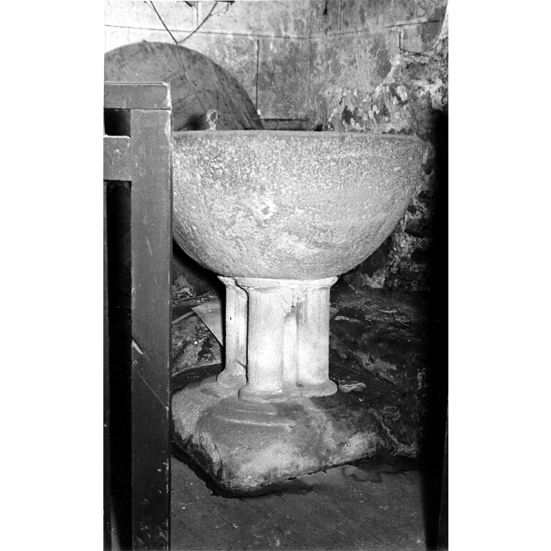 Source caption: "Pila bautismal de la iglesia de San Juan de Oyardo [...] pieza medieval con la copa lisa. Se apea en un pie con cuatro medias columnas adosadas a un núcleo central que se apoya en una basa cuadrada con decoración de garras" [NB: the round flat cover stands against the wall behind the font]