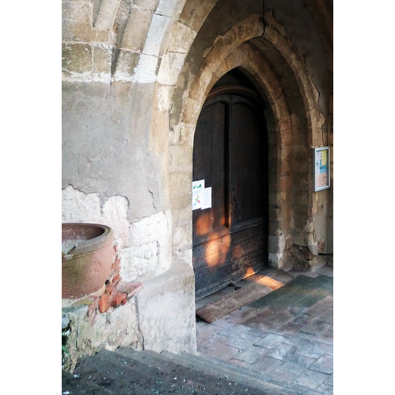 Source caption: "Porche de l'église Saint-Barthélemy" -- the reddish vessel on the left, sitting on a stone ledge of the porch, is quite likely the remains of a baptismal font