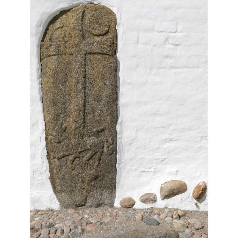 "romansk, af grovkornet grå og rød granit, synlig højde 162 cm, bredde ved korsarmen 65 cm, ved jorden 55 cm. Kraftigt reliefhugget, latinsk kors, hvis øvre arme flankeres af sol og måne, og hvis stamme hviler på ryggen af en løve. Denne har ret lang snude og hængende tunge samt lang hale endende i trebladet dusk; hele motivet svarer nøje til det på en sten i den nærliggende Skødstrup kirke (Randers amt). Indmuret i våbenhusets østmur, hvor den også nævnes i 1600-tallet." [source: DK]